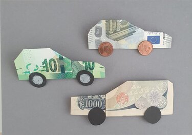 Drei Origami-Autos aus Banknoten mit Münz- und Papierrädern auf grauem Hintergrund.