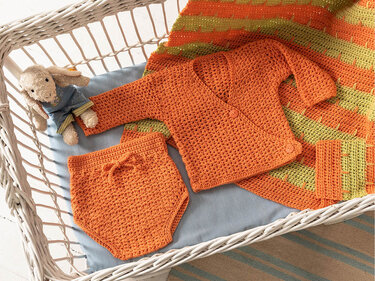 Orangefarbenes gehäkeltes Babyset aus Wickeljacke mit Knöpfen und kurzer Hose in einer weißen Korbwiege, daneben Plüschhase und gestreifte Häkeldecke.