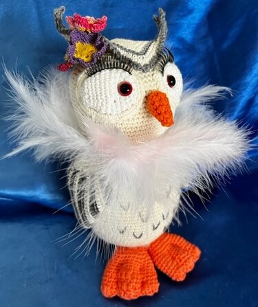 Gehäkelte weiße Amigurumi-Eule mit flauschigem weißem Federkragen, orangefarbenem Schnabel und Füßen, roten Augen, bunter Blume am Kopf