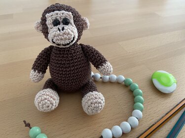 Kleiner gehäkelter Amigurumi-Affe in Braun und Beige auf Holztisch neben Perlenkette, Radierer und Stiften