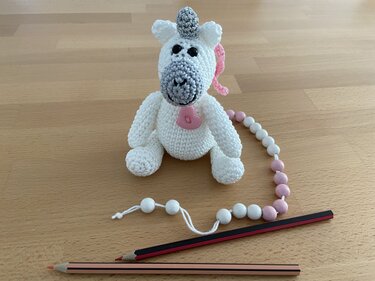 Weißes Einhorn-Amigurumi mit rosa Mähne und Perlenkette auf Holzoberfläche neben zwei Buntstiften