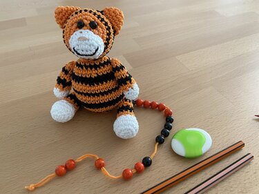 Gehäkeltes Tiger-Amigurumi in Orange-Schwarz mit weißer Schnauze und Pfoten, daneben eine Perlenkette, zwei Stifte und ein Radiergummi