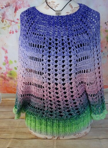 Vorderansicht eines gehäkelten Ponchos mit lila-rosa-grünem Farbverlauf, Lochmuster und geripptem Saum auf einer Schneiderpuppe