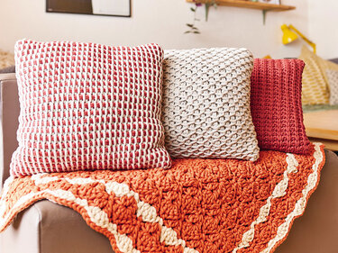 Nahaufnahme von drei gehäkelten Kissen in Rot, Weiß und Beige sowie einer orangefarbenen Häkeldecke auf einem Sofa.