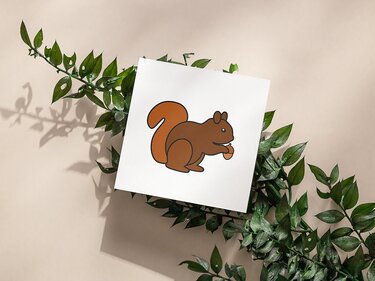 Braunes, stilisiertes Eichhörnchen hält eine Nuss auf einem quadratischen weißen Blatt, arrangiert auf grünen Blättern.