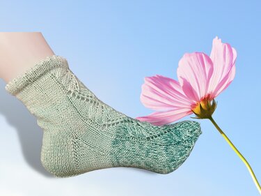 Pastellgrüne gestrickte Kindersocke mit Rüschenbündchen und Blatt-Ajourkante, seitliche Ansicht neben rosa Blüte vor hellem Himmel