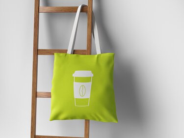 Grüne Stofftasche mit weißem Kaffeebecher-Symbol und Kaffeebohne an einer Holzleiter.