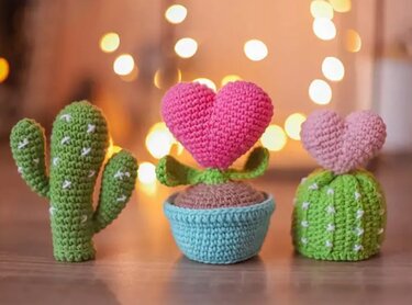 Drei gehäkelte Mini‑Kakteen und ein Topf mit herzförmiger Blüte in Pastellfarben, vor unscharfen Lichtern arrangiert