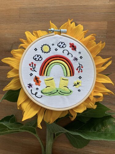 Stickrahmen mit gesticktem Regenbogen, gelben Gummistiefeln, Sonne, Wolken und Herbstblättern auf einer Sonnenblume