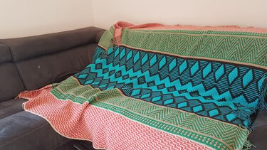 Mehrfarbige gehäkelte Decke mit türkisen, grünen und rosa geometrischen Mustern auf dunklem Sofa