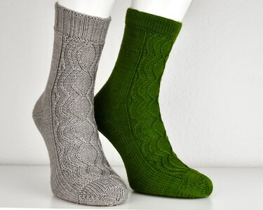 Zwei gestrickte Socken auf weißen Fußformen; eine grau, eine dunkelgrün, beide mit Zopfmuster.