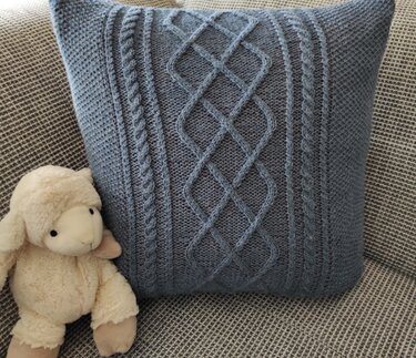 Blaugrauer gestrickter Kissenbezug mit Zopfmuster auf Sofa neben Stofflamm