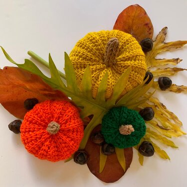 Three crocheted pumpkins in orange, yellow, and green arranged with fall leaves and seed pods on a light background