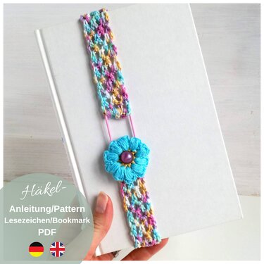 Hand holding white hardcover book with a multicolored crocheted strap bookmark and a turquoise flower attached by an elastic loop