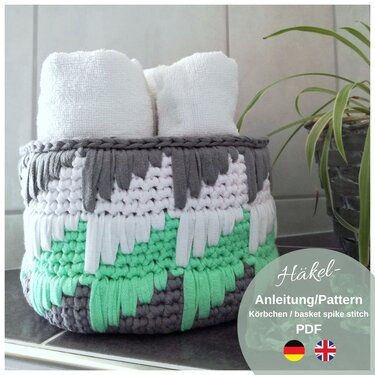 Round crochet basket in gray, white and mint with spike-stitch fringe, holding two rolled white towels on a tiled bathroom surface beside a potted plant