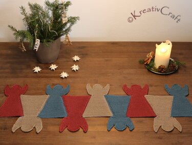 Crocheted angel-motif table runner in red, blue and beige on a wooden table with candle and small pine