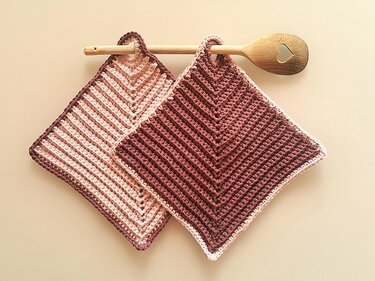 Zwei gehäkelte quadratische Topflappen in hellen und dunklen Brauntönen mit V‑förmigem Muster an einem Holzlöffel hängend