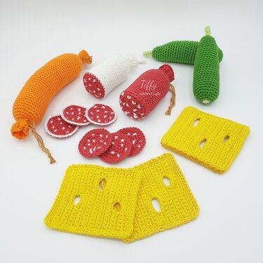 Crocheted toy groceries: yellow cheese slices, red salami rounds, orange and white sausages, and green cucumbers on a white background
