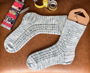 Zwei blau-weiß gestrickte Socken mit Lochmuster auf braunem Untergrund, daneben Stricknadeln und Maßband.