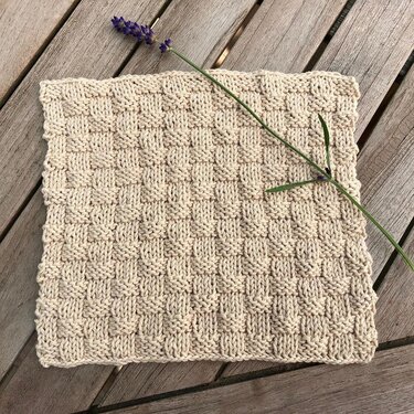 Beiger handgestrickter Topflappen mit Korbmuster und Lavendelzweig auf Holz.