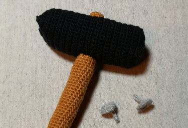Gehäkelter Hammer mit schwarzem Kopf und braunem Stiel neben zwei kleinen gehäkelten Nägeln auf hellem Hintergrund.