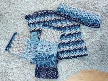 Gehäkeltes Set aus Schal, fingerlosen Handschuhen und Stirnband in Blau-, Weiß- und Grautönen auf einem flauschigen Untergrund.
