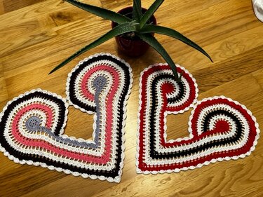 Zwei gehäkelte Herz-Deckchen in Rot, Rosa, Schwarz und Creme auf Holztisch, teils verdeckt von einer Zimmerpflanze