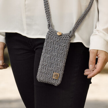 Grau gestrickte Handytasche mit Umhängeband und Holzknopf, getragen über einer weißen Bluse und schwarzen Hosen.