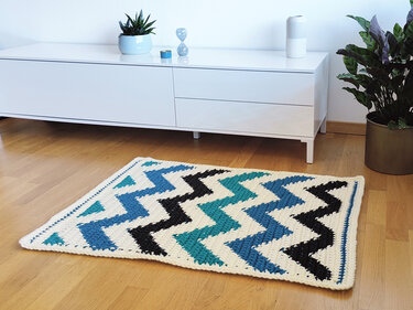 Gehäkelter Teppich in Creme, Blau, Türkis und Schwarz mit Zickzack-Muster auf Holzboden vor weißem Sideboard