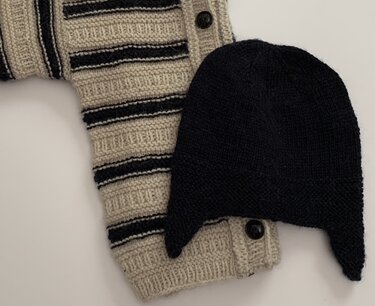 Gestrickte beigefarbene Babyjacke mit dunklen Streifen neben einer dunklen Babymütze