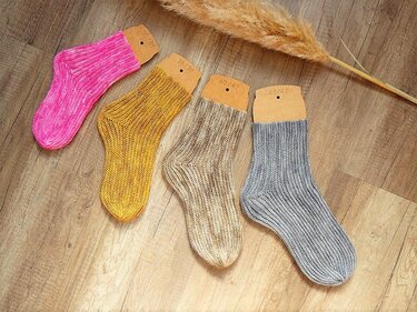 Vier handgehäkelte Rippensocken in Pink, Gelb, Beige und Grau auf Holzboden mit Karton-Einlegern.