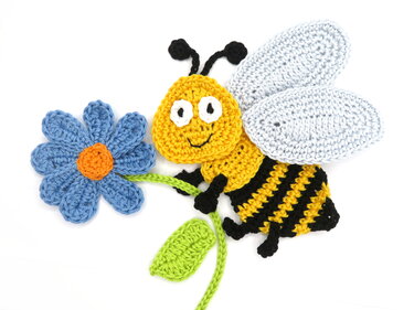 Flat crochet bee applique in yellow and black holding a blue crochet flower on white background