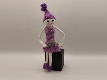 Gehäkelte Amigurumi-Puppe in Lila mit Bommelmütze, sitzend auf schwarzem Würfel, hält kleines Garnprojekt.