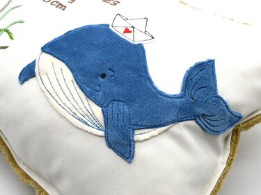 Gestickter blauer Wal mit kleinem Papierschiff als Applikation auf einem hellen Kissen