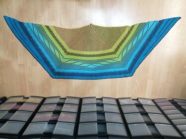Halbrundes, gestricktes Tuch in Blau und Grün mit schwarzen Linien und geometrischem Muster auf hellem Holzfußboden ausgebreitet.