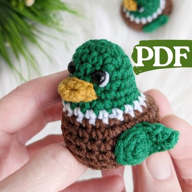 Hand holding a tiny crocheted amigurumi bird with green head, yellow beak, brown body and leaf-shaped wing; PDF overlay in corner