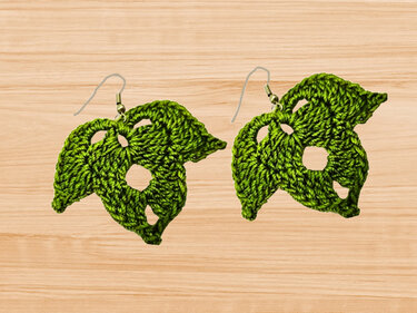 Pair of green crocheted Monstera leaf earrings with silver-tone hooks on a light wooden background.