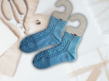 Nahaufnahme handgestrickter blauer Knöchelsocken mit geripptem Bündchen und Zopfmuster neben Garn und Schere