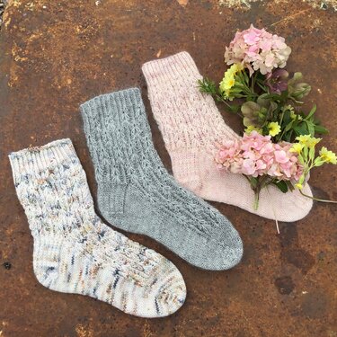 Drei gestrickte Damensocken in Rosa, Grau und meliertem Garn mit erkennbaren Musterdetails und Blumenarrangement