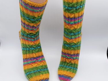 Wadenlange, bunte gestrickte Socken mit Zopf- und Querstreifen in Gelb, Grün, Blau und Pink an Beinen vor weißem Hintergrund.