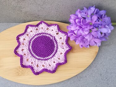 lavender-toned crochet coaster with scalloped star edge next to purple artificial flower on wooden board