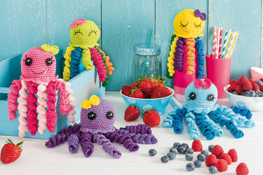 Fünf gehäkelte Oktopusse in Pink, Grün, Lila, Gelb und Blau mit Schalen voller Erdbeeren, Himbeeren und Blaubeeren vor einer türkisfarbenen Holzwand.