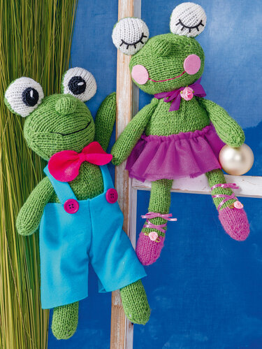 Zwei gestrickte grüne Amigurumi-Frösche, einer mit blauer Latzhose und Fliege, der andere mit violettem Tüllrock und Schlafaugen.