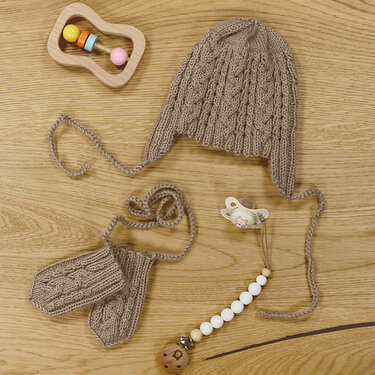 Beige handgestrickte Babymütze mit Ohrenklappen und passende Fäustlinge im Zopfmuster auf Holzuntergrund, daneben Holzrassel und Schnuller.