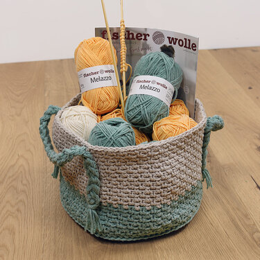 Zweifarbiger gehäkelter Korb in Beige und Mintgrün, gefüllt mit Wollknäueln und Stricknadeln.