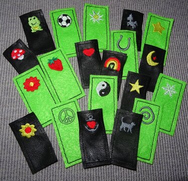 ITH-Stickanhänger aus grünem Filz und schwarzem Kunstleder mit Frosch-, Fußball-, Herz- und Yin‑Yang-Motiven auf grauem Untergrund.