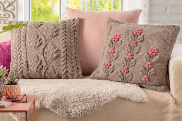 Gestrickte Kissen mit Zopf- und Blattmuster sowie rosa Blüten auf Sofa