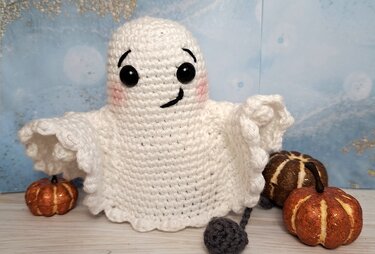 Weißes gehäkeltes Amigurumi-Gespenst mit schwarzen Augen und rosa Wangen, umgeben von kleinen Kürbissen
