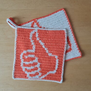 Zwei gehäkelte quadratische Topflappen in Orange und Weiß mit Daumen‑hoch‑Motiv auf Holzuntergrund.