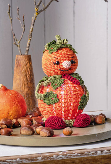 Gehäkeltes Kürbismännchen mit transparentem Kugelbauch, grünen Blättern und roten Häkel-Schuhen auf einem Tablett mit Kastanien.